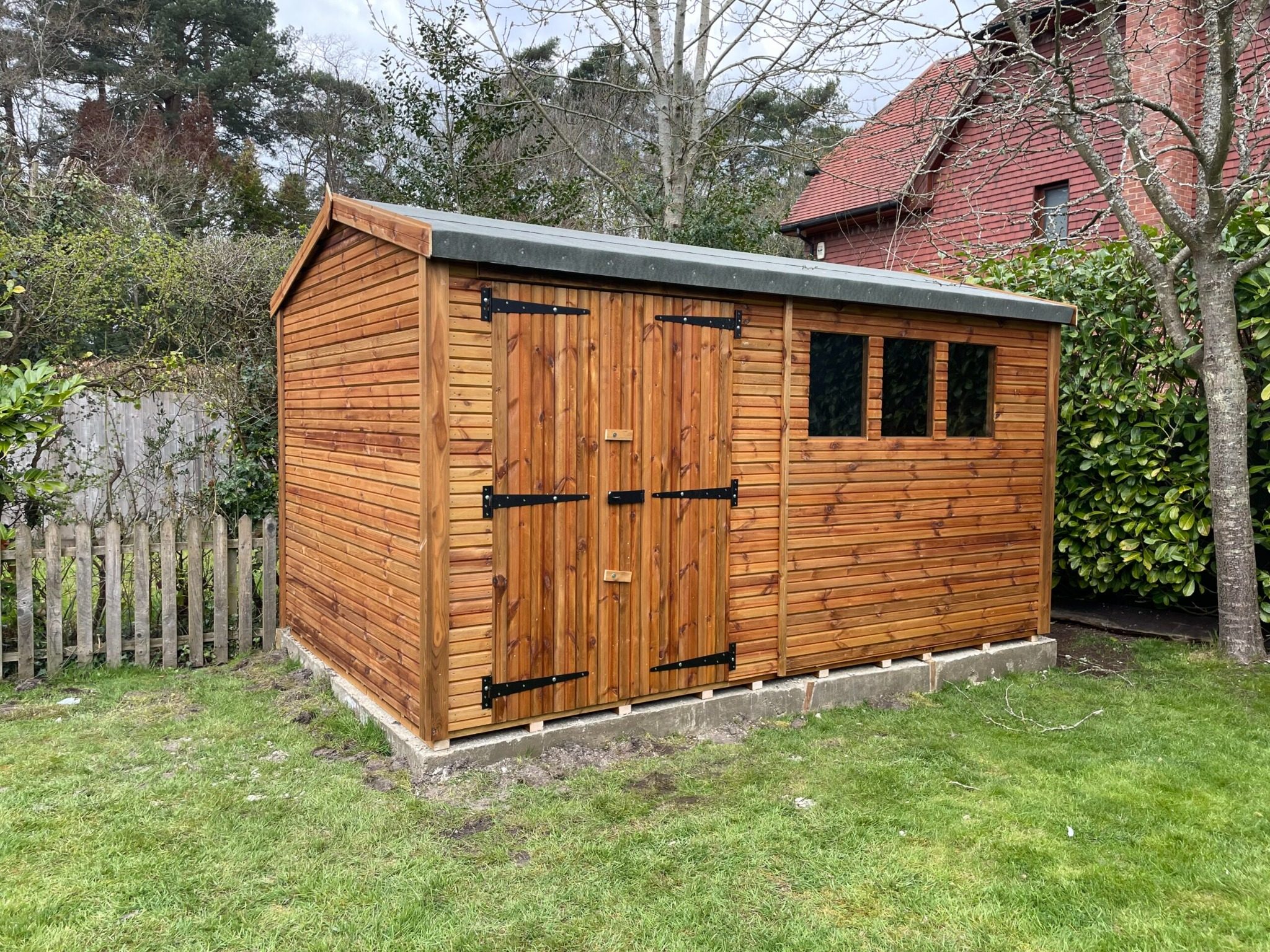 Large wooden garden shed with double doors, three side windows, and a felted apex roof, installed on a concrete base in a residential garden.